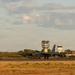 A large military bomber aircraft, identifiable as a B-52 Stratofortress, is taxiing or stationary on a runway at a military airfield. Two air traffic control towers are visible in the background, along with what appears to be a hardened aircraft shelter or hangar to the right. The image is taken during golden hour lighting conditions, suggesting early morning or late afternoon.