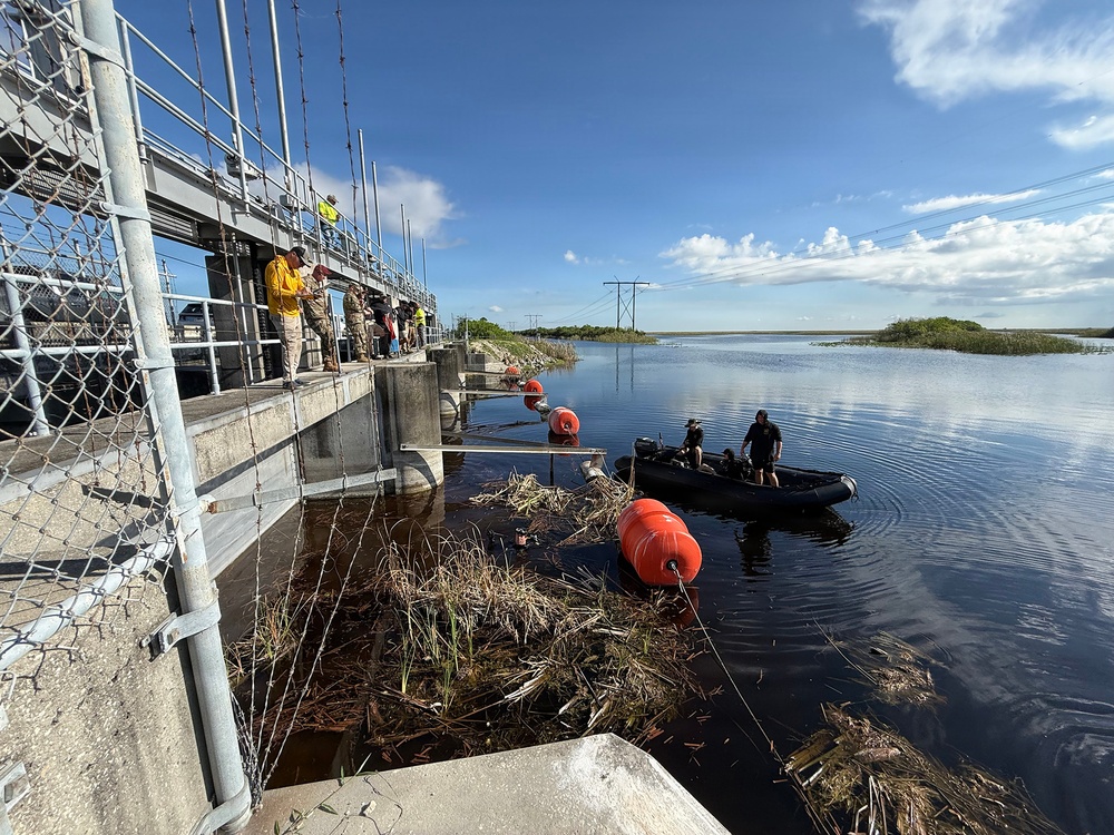 The image shows a water control structure or flood gate facility where personnel are conducting operations involving a black inflatable boat and orange marker buoys on a calm waterway. Several individuals are visible on the concrete structure/walkway observing or directing activities, while two people operate the boat near accumulated aquatic debris. The setting appears to be a wetland or managed water system, possibly in Florida based on the vegetation and landscape.