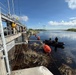 The image shows a water control structure or flood gate facility where personnel are conducting operations involving a black inflatable boat and orange marker buoys on a calm waterway. Several individuals are visible on the concrete structure/walkway observing or directing activities, while two people operate the boat near accumulated aquatic debris. The setting appears to be a wetland or managed water system, possibly in Florida based on the vegetation and landscape.