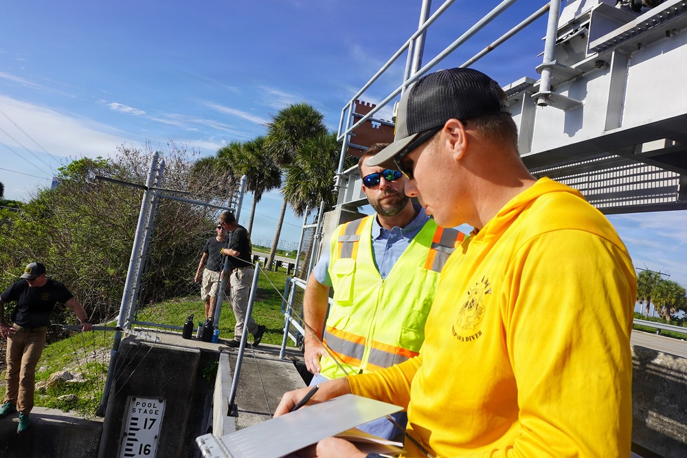 A group of workers or inspectors are gathered at what appears to be a water control structure or flood gate facility. Two men in the foreground are reviewing documents on a clipboard, with one wearing a yellow hoodie and a cap, and the other wearing a high-visibility safety vest. Additional personnel are visible in the background near a chain-link fence.