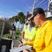 A group of workers or inspectors are gathered at what appears to be a water control structure or flood gate facility. Two men in the foreground are reviewing documents on a clipboard, with one wearing a yellow hoodie and a cap, and the other wearing a high-visibility safety vest. Additional personnel are visible in the background near a chain-link fence.