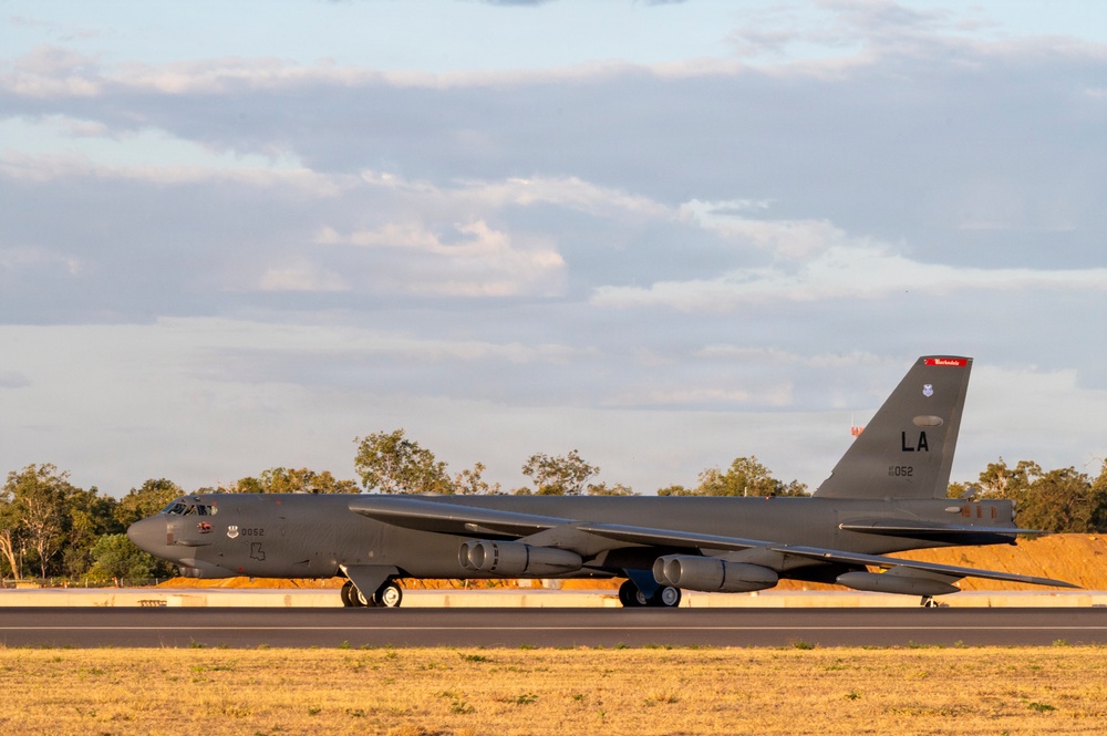 A U.S. Air Force B-52 Stratofortress strategic bomber is taxiing on a runway, identifiable by the tail code 'LA' and serial number ending in '052'. The aircraft is painted in standard grey livery and appears to be at an airfield surrounded by eucalyptus-type trees, suggesting a location in Australia. The image was taken during golden hour lighting conditions, casting warm tones across the scene.