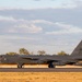 A U.S. Air Force B-52 Stratofortress strategic bomber is taxiing on a runway, identifiable by the tail code 'LA' and serial number ending in '052'. The aircraft is painted in standard grey livery and appears to be at an airfield surrounded by eucalyptus-type trees, suggesting a location in Australia. The image was taken during golden hour lighting conditions, casting warm tones across the scene.