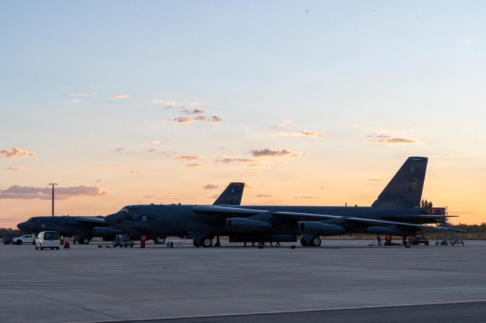 Two B-52 Stratofortress strategic bombers are parked on a flight line apron at dusk or dawn, with ground crews performing maintenance or pre/post-flight operations. The aircraft display 'LA' tail codes, indicating assignment to Barksdale Air Force Base, Louisiana (2nd Bomb Wing). The warm sunset sky provides a dramatic backdrop to the military flight line scene.