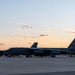 Two B-52 Stratofortress strategic bombers are parked on a flight line apron at dusk or dawn, with ground crews performing maintenance or pre/post-flight operations. The aircraft display 'LA' tail codes, indicating assignment to Barksdale Air Force Base, Louisiana (2nd Bomb Wing). The warm sunset sky provides a dramatic backdrop to the military flight line scene.