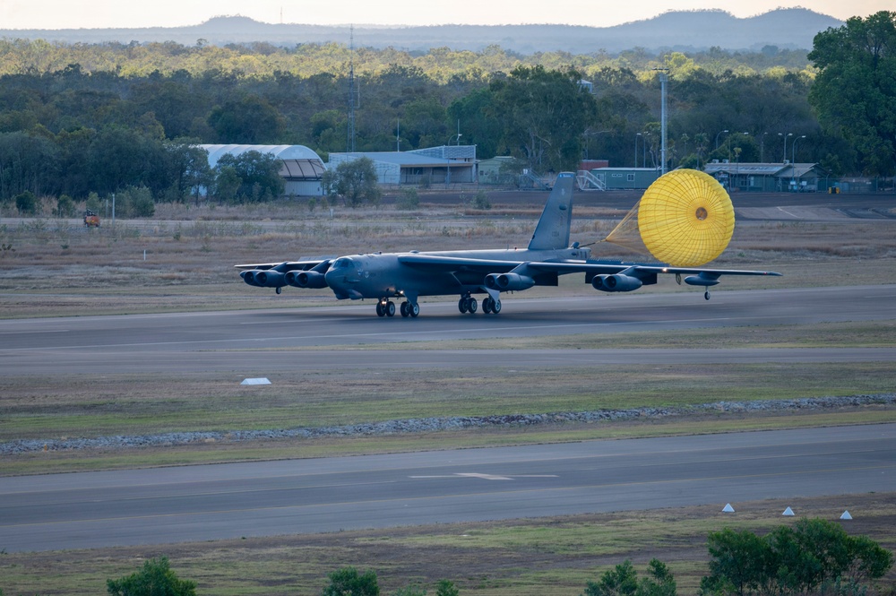 A large military bomber aircraft, identifiable as a B-52 Stratofortress, is landing on a runway with a bright yellow drag parachute deployed behind it to assist in deceleration. The aircraft is on the ground with all landing gear visible, rolling down the tarmac. The setting appears to be a military airfield surrounded by sparse vegetation, low buildings, and hills in the background, consistent with an Australian location.