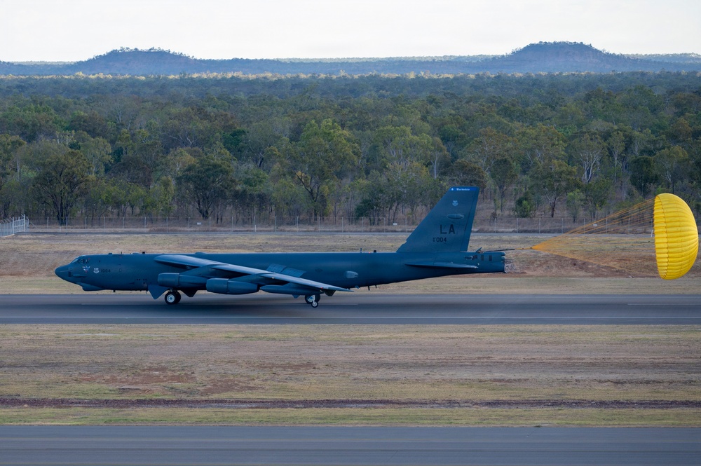 A U.S. Air Force B-52 Stratofortress strategic bomber is landing on a runway with its yellow drag parachute deployed to assist braking. The aircraft bears the tail code 'LA' and serial number '1004', indicating it belongs to the 2nd Bomb Wing based at Barksdale Air Force Base, Louisiana. The setting appears to be a military airfield surrounded by Australian eucalyptus woodland and low mesa-like hills in the background.