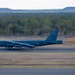 A U.S. Air Force B-52 Stratofortress strategic bomber is landing on a runway with its yellow drag parachute deployed to assist braking. The aircraft bears the tail code 'LA' and serial number '1004', indicating it belongs to the 2nd Bomb Wing based at Barksdale Air Force Base, Louisiana. The setting appears to be a military airfield surrounded by Australian eucalyptus woodland and low mesa-like hills in the background.