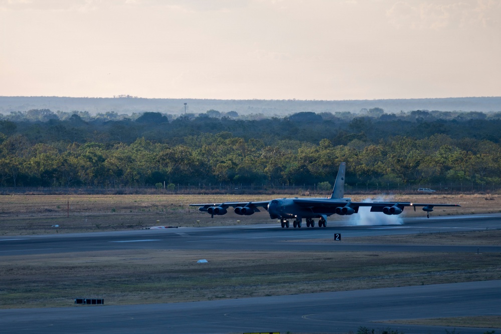 A B-52 Stratofortress strategic bomber is landing or rolling out on a runway, with visible smoke or steam from the braking system beneath the aircraft. The image is taken at dusk or dawn in low-light conditions, with a treeline and rolling terrain visible in the background. A runway marker with the number '2' is visible in the foreground.