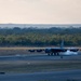A B-52 Stratofortress strategic bomber is landing or rolling out on a runway, with visible smoke or steam from the braking system beneath the aircraft. The image is taken at dusk or dawn in low-light conditions, with a treeline and rolling terrain visible in the background. A runway marker with the number '2' is visible in the foreground.