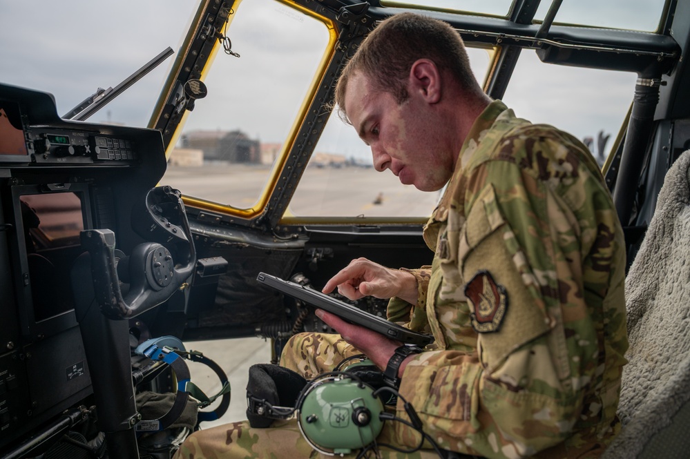 A U.S. Air Force service member in OCP (Operational Camouflage Pattern) uniform is seated in what appears to be a military aircraft cockpit, operating a ruggedized tablet device. The individual is focused on the tablet screen while surrounded by cockpit instrumentation and controls. The setting appears to be on a flight line or airfield, visible through the open cockpit window.