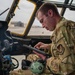 A U.S. Air Force service member in OCP (Operational Camouflage Pattern) uniform is seated in what appears to be a military aircraft cockpit, operating a ruggedized tablet device. The individual is focused on the tablet screen while surrounded by cockpit instrumentation and controls. The setting appears to be on a flight line or airfield, visible through the open cockpit window.