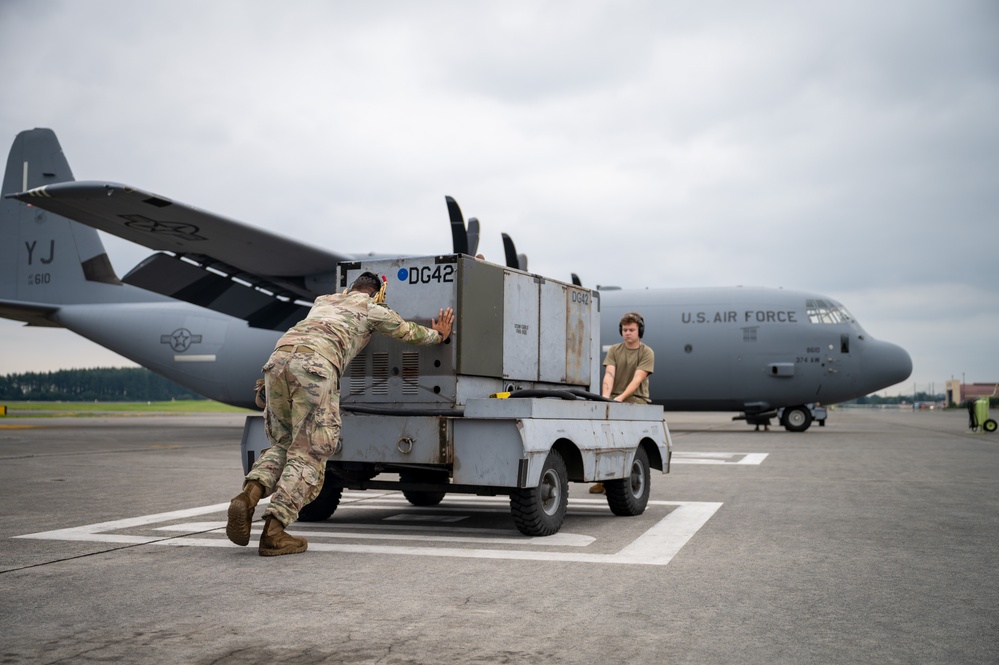 Two U.S. Air Force personnel are working on a flight line, pushing ground support equipment on a wheeled cart in front of a C-130 Hercules transport aircraft. The aircraft bears the tail code 'YJ' and serial number ending in '610', with 'U.S. Air Force' markings visible on the fuselage. The scene appears to be at a military air base under overcast skies.