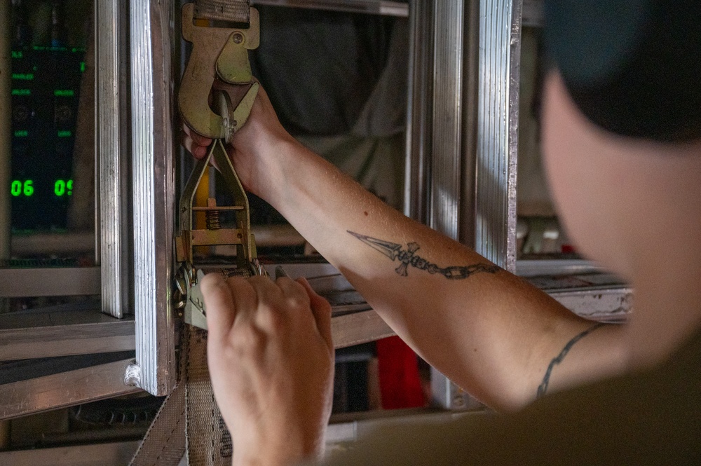 A person is working with a ratchet strap and metal hook mechanism, securing or tensioning cargo within what appears to be a military or industrial transport environment. The individual has a visible tattoo on their forearm depicting a cross with a chain or arrow design. Green digital displays are visible in the background, suggesting an aircraft or vehicle cargo bay setting.