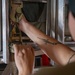 A person is working with a ratchet strap and metal hook mechanism, securing or tensioning cargo within what appears to be a military or industrial transport environment. The individual has a visible tattoo on their forearm depicting a cross with a chain or arrow design. Green digital displays are visible in the background, suggesting an aircraft or vehicle cargo bay setting.