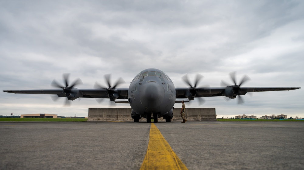 A large four-engine turboprop military transport aircraft, consistent with a C-130 Hercules variant, is photographed head-on on a tarmac with all four propellers spinning (motion blur visible). A single uniformed military personnel stands in front of the aircraft near the nose gear, likely conducting a pre-flight or post-flight inspection. The image is taken from a low angle along a yellow centerline marking, with a concrete blast deflector wall visible behind the aircraft's landing gear.