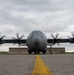 A large four-engine turboprop military transport aircraft, consistent with a C-130 Hercules variant, is photographed head-on on a tarmac with all four propellers spinning (motion blur visible). A single uniformed military personnel stands in front of the aircraft near the nose gear, likely conducting a pre-flight or post-flight inspection. The image is taken from a low angle along a yellow centerline marking, with a concrete blast deflector wall visible behind the aircraft's landing gear.