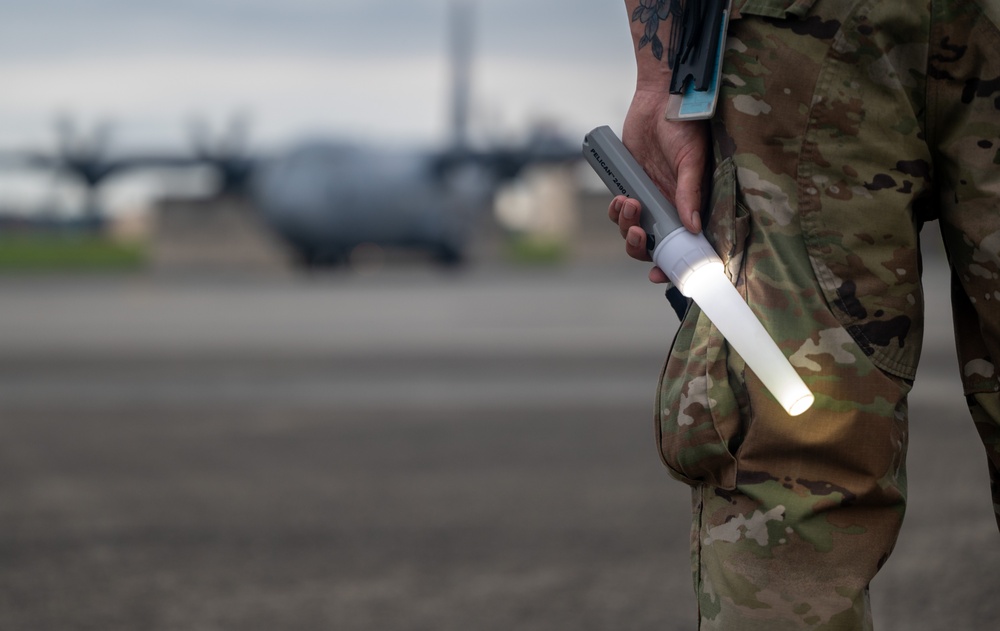 A military service member in MultiCam/OCP camouflage uniform stands on an airfield tarmac holding an illuminated Pelican flashlight with a cone/wand attachment. The light wand is glowing white, suggesting low-light or nighttime operations. In the blurred background, at least one C-130 Hercules military transport aircraft is visible on the flight line.