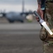 A military service member in MultiCam/OCP camouflage uniform stands on an airfield tarmac holding an illuminated Pelican flashlight with a cone/wand attachment. The light wand is glowing white, suggesting low-light or nighttime operations. In the blurred background, at least one C-130 Hercules military transport aircraft is visible on the flight line.