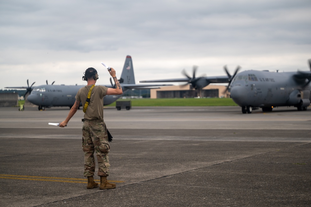 A U.S. Air Force ground crew member is marshaling military transport aircraft on a flight line using signal wands. Two C-130J Super Hercules aircraft marked 'U.S. AIR FORCE' are visible on the tarmac with their propellers spinning. The scene appears to take place at a military airfield under overcast skies.