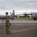 A U.S. Air Force ground crew member is marshaling military transport aircraft on a flight line using signal wands. Two C-130J Super Hercules aircraft marked 'U.S. AIR FORCE' are visible on the tarmac with their propellers spinning. The scene appears to take place at a military airfield under overcast skies.