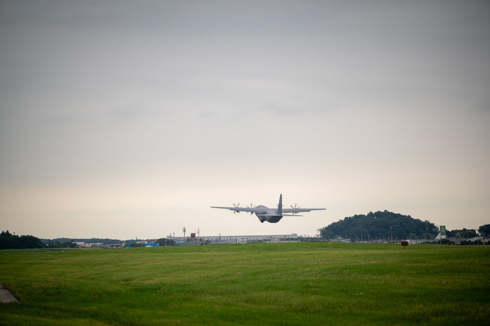A military turboprop transport aircraft, consistent with a C-130 Hercules configuration, is captured at low altitude during takeoff or landing over a grass airfield. The overcast sky and lush green terrain suggest a temperate, possibly East Asian location based on the visible infrastructure and vegetation. Industrial or base facilities and a tree-covered hill are visible in the background.