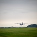 A military turboprop transport aircraft, consistent with a C-130 Hercules configuration, is captured at low altitude during takeoff or landing over a grass airfield. The overcast sky and lush green terrain suggest a temperate, possibly East Asian location based on the visible infrastructure and vegetation. Industrial or base facilities and a tree-covered hill are visible in the background.