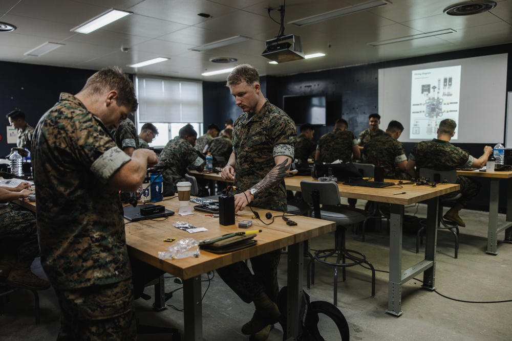 A group of military personnel in camouflage uniforms are engaged in a hands-on technical training session in a classroom environment. Participants are working at wooden workbenches with various electronic components and tools. A projected presentation displaying what appears to be a wiring diagram is visible on the screen at the front of the room.