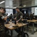 A group of military personnel in camouflage uniforms are engaged in a hands-on technical training session in a classroom environment. Participants are working at wooden workbenches with various electronic components and tools. A projected presentation displaying what appears to be a wiring diagram is visible on the screen at the front of the room.