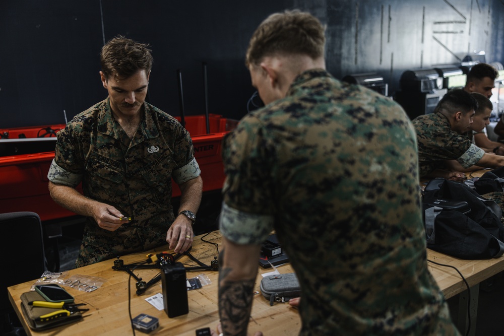 Military personnel in MARPAT (Marine pattern) camouflage uniforms are working at a workbench in what appears to be a technical workshop or lab environment. The individuals appear to be assembling or maintaining small drone/UAV components and electronic equipment. Multiple service members are visible, with some working at computers in the background.