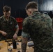 Military personnel in MARPAT (Marine pattern) camouflage uniforms are working at a workbench in what appears to be a technical workshop or lab environment. The individuals appear to be assembling or maintaining small drone/UAV components and electronic equipment. Multiple service members are visible, with some working at computers in the background.
