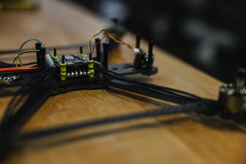 The image shows a partially assembled FPV (First Person View) racing drone frame resting on a wooden surface. A flight controller board is mounted at the center of the carbon fiber frame, with multiple colored wires routed along the arms. A brushless motor is partially visible on the right arm of the frame.
