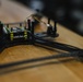 The image shows a partially assembled FPV (First Person View) racing drone frame resting on a wooden surface. A flight controller board is mounted at the center of the carbon fiber frame, with multiple colored wires routed along the arms. A brushless motor is partially visible on the right arm of the frame.