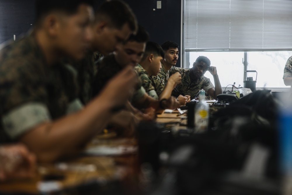A group of military personnel in camouflage uniforms are seated at a long table in what appears to be a briefing room or classroom setting. The individuals are engaged in what looks like a training session, review, or planning meeting. The image uses a shallow depth of field, with foreground figures blurred and focus on mid-ground subjects.
