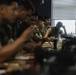 A group of military personnel in camouflage uniforms are seated at a long table in what appears to be a briefing room or classroom setting. The individuals are engaged in what looks like a training session, review, or planning meeting. The image uses a shallow depth of field, with foreground figures blurred and focus on mid-ground subjects.