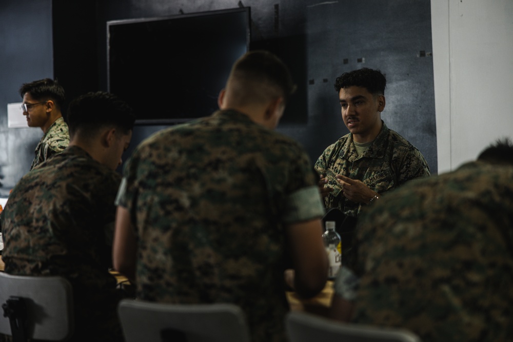A group of U.S. Marines in woodland MARPAT camouflage uniforms are gathered around a table in what appears to be an indoor briefing or meeting room. One Marine facing the camera appears to be speaking or explaining something to the others. The setting suggests a planning session, briefing, or informal discussion.