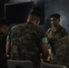A group of U.S. Marines in woodland MARPAT camouflage uniforms are gathered around a table in what appears to be an indoor briefing or meeting room. One Marine facing the camera appears to be speaking or explaining something to the others. The setting suggests a planning session, briefing, or informal discussion.
