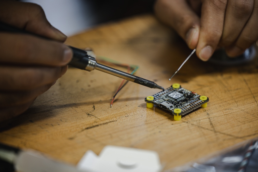 A person is soldering wires onto a small printed circuit board (PCB) placed on a wooden workbench. The PCB features yellow corner standoffs and a central microcontroller chip, consistent with a flight controller or similar embedded electronics module. The work appears to be hands-on electronics assembly or repair.