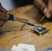 A person is soldering wires onto a small printed circuit board (PCB) placed on a wooden workbench. The PCB features yellow corner standoffs and a central microcontroller chip, consistent with a flight controller or similar embedded electronics module. The work appears to be hands-on electronics assembly or repair.