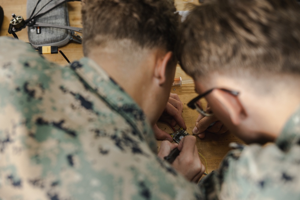 Two individuals wearing US Marine Corps MARPAT digital camouflage uniforms are closely examining and working on a small electronic circuit board on a wooden surface. One individual is wearing glasses and both are using tools, appearing to be soldering or assembling electronics. In the background, what appears to be a small drone or similar device is visible on the table.