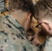 Two individuals wearing US Marine Corps MARPAT digital camouflage uniforms are closely examining and working on a small electronic circuit board on a wooden surface. One individual is wearing glasses and both are using tools, appearing to be soldering or assembling electronics. In the background, what appears to be a small drone or similar device is visible on the table.