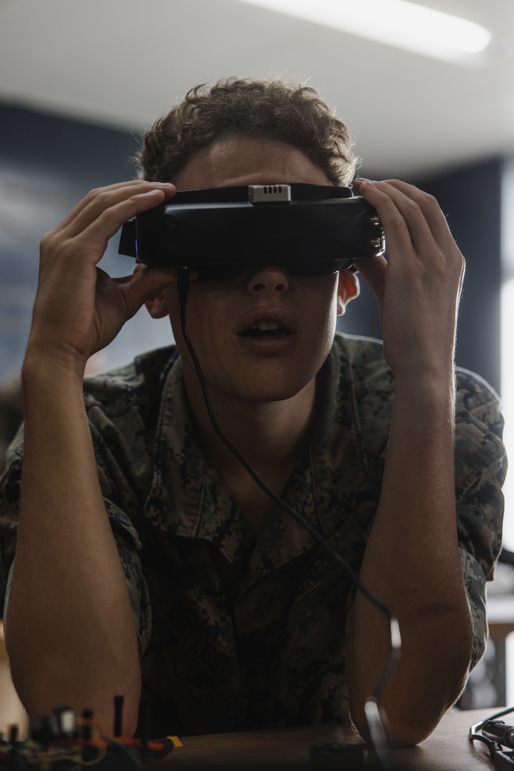 A young person wearing military-style camouflage clothing is holding a VR or FPV headset up to their eyes with both hands. The setting appears to be an indoor classroom or training environment with fluorescent lighting. Electronic components or a controller are visible on the table in front of them.