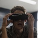 A young person wearing military-style camouflage clothing is holding a VR or FPV headset up to their eyes with both hands. The setting appears to be an indoor classroom or training environment with fluorescent lighting. Electronic components or a controller are visible on the table in front of them.