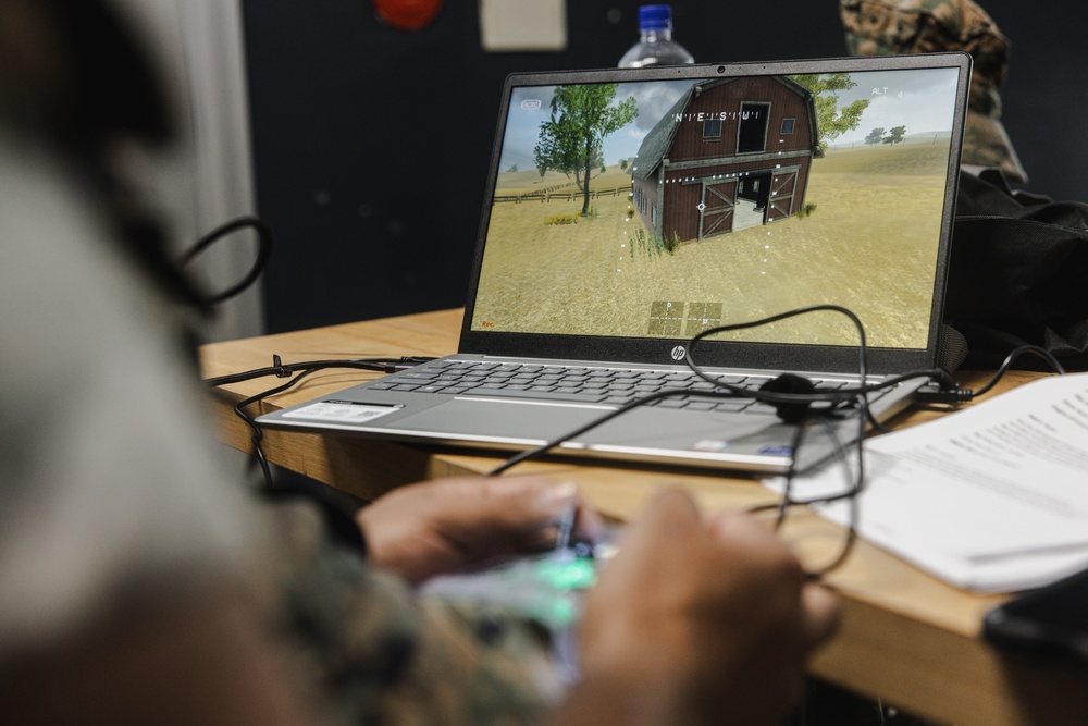 A person in military camouflage uniform is operating what appears to be a drone or simulation controller while viewing a 3D virtual environment on an HP laptop. The screen displays a first-person or aerial perspective of a rural scene featuring a red barn, open fields, and trees, consistent with military simulation or drone training software. Papers and cables are visible on the wooden desk alongside the laptop.
