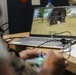 A person in military camouflage uniform is operating what appears to be a drone or simulation controller while viewing a 3D virtual environment on an HP laptop. The screen displays a first-person or aerial perspective of a rural scene featuring a red barn, open fields, and trees, consistent with military simulation or drone training software. Papers and cables are visible on the wooden desk alongside the laptop.
