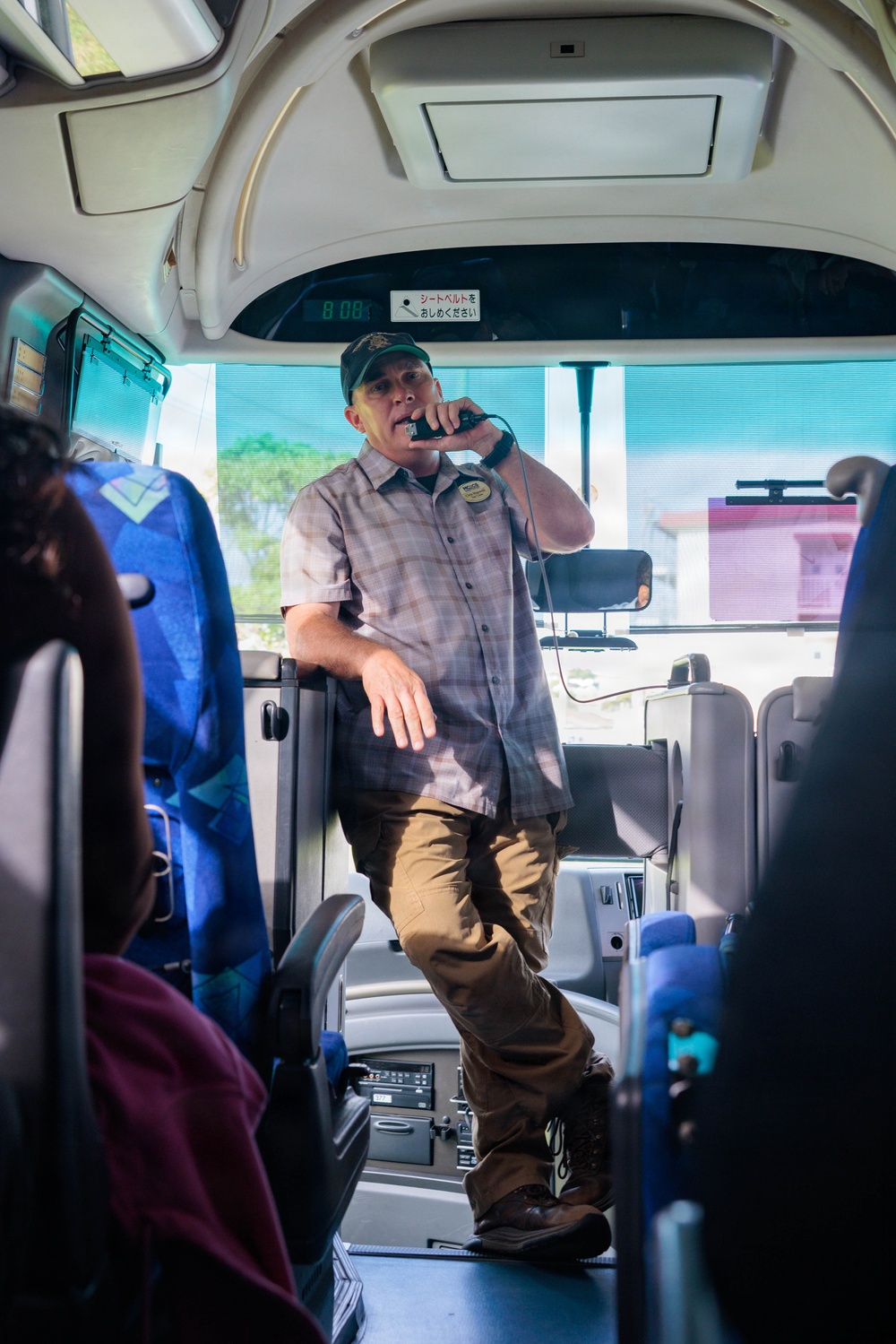 A man wearing a green cap and plaid shirt stands at the front of a tour or charter bus, speaking into a handheld microphone. He appears to be a tour guide or bus staff member addressing passengers. Japanese text is visible on a sign above him, and the bus interior features blue patterned seats.