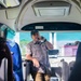 A man wearing a green cap and plaid shirt stands at the front of a tour or charter bus, speaking into a handheld microphone. He appears to be a tour guide or bus staff member addressing passengers. Japanese text is visible on a sign above him, and the bus interior features blue patterned seats.
