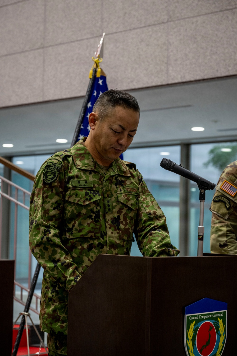 A military officer in Japan Ground Self-Defense Force (JGSDF) camouflage uniform stands at a podium bearing the 'Ground Component Command' insignia. He appears to be delivering a speech or reading remarks at a formal military ceremony. A U.S. flag and a partially visible U.S. Army soldier are present in the background, indicating a joint bilateral military event.