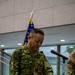 A military officer in Japan Ground Self-Defense Force (JGSDF) camouflage uniform stands at a podium bearing the 'Ground Component Command' insignia. He appears to be delivering a speech or reading remarks at a formal military ceremony. A U.S. flag and a partially visible U.S. Army soldier are present in the background, indicating a joint bilateral military event.