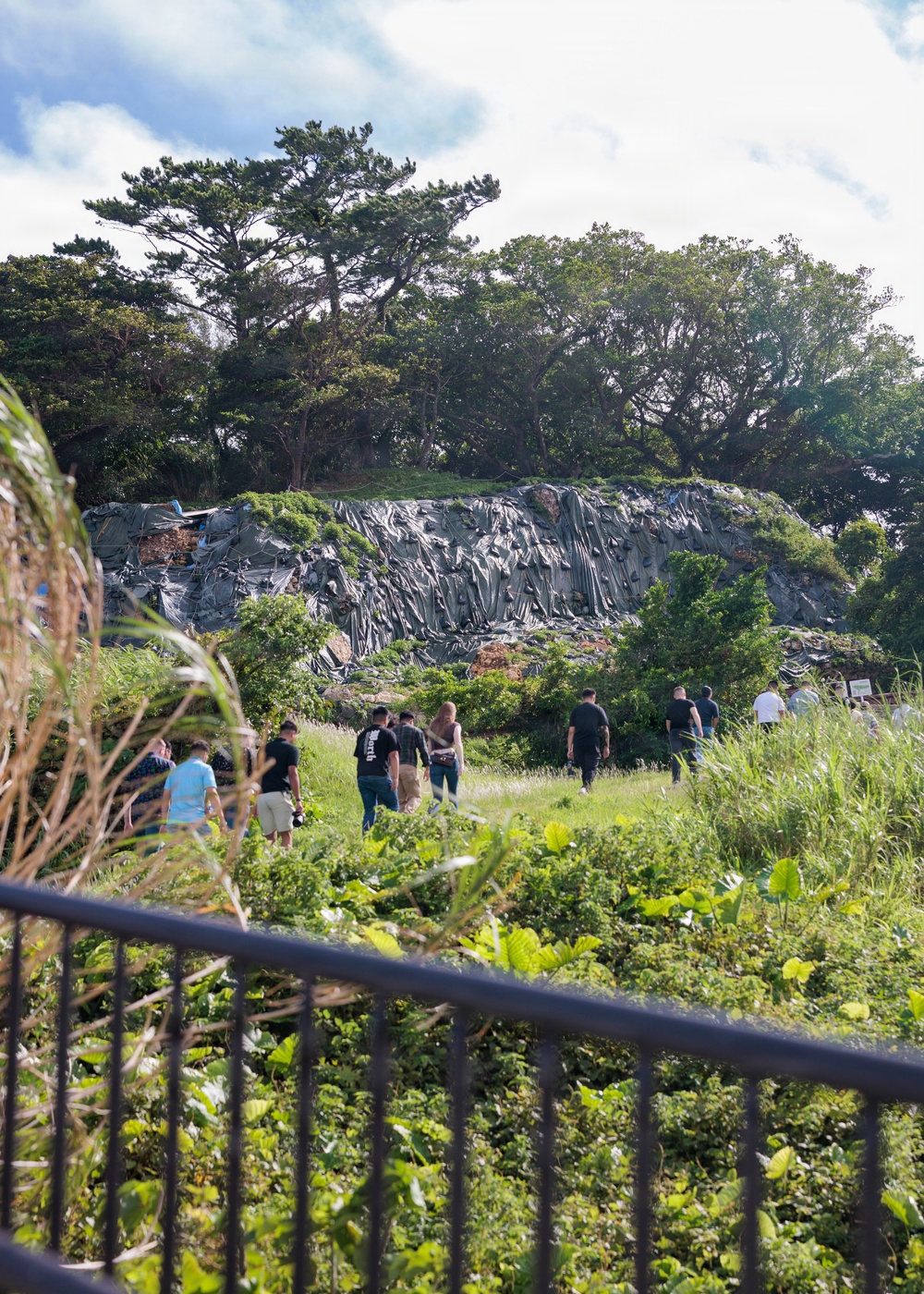 A group of approximately 8-10 people are walking through a grassy, overgrown area toward a large rocky cliff face covered with dark, layered geological formations or possibly tarpaulin/sheeting material. The scene is viewed through a metal railing in the foreground, with lush tropical vegetation surrounding the path. Large trees crown the top of the rocky formation under a partly cloudy sky.